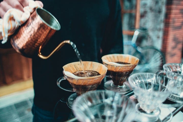 A person pours hot water from a copper kettle into a pour-over coffee filter, brewing coffee. Several glass coffee drippers are arranged on a metal rack. The background is slightly blurred.