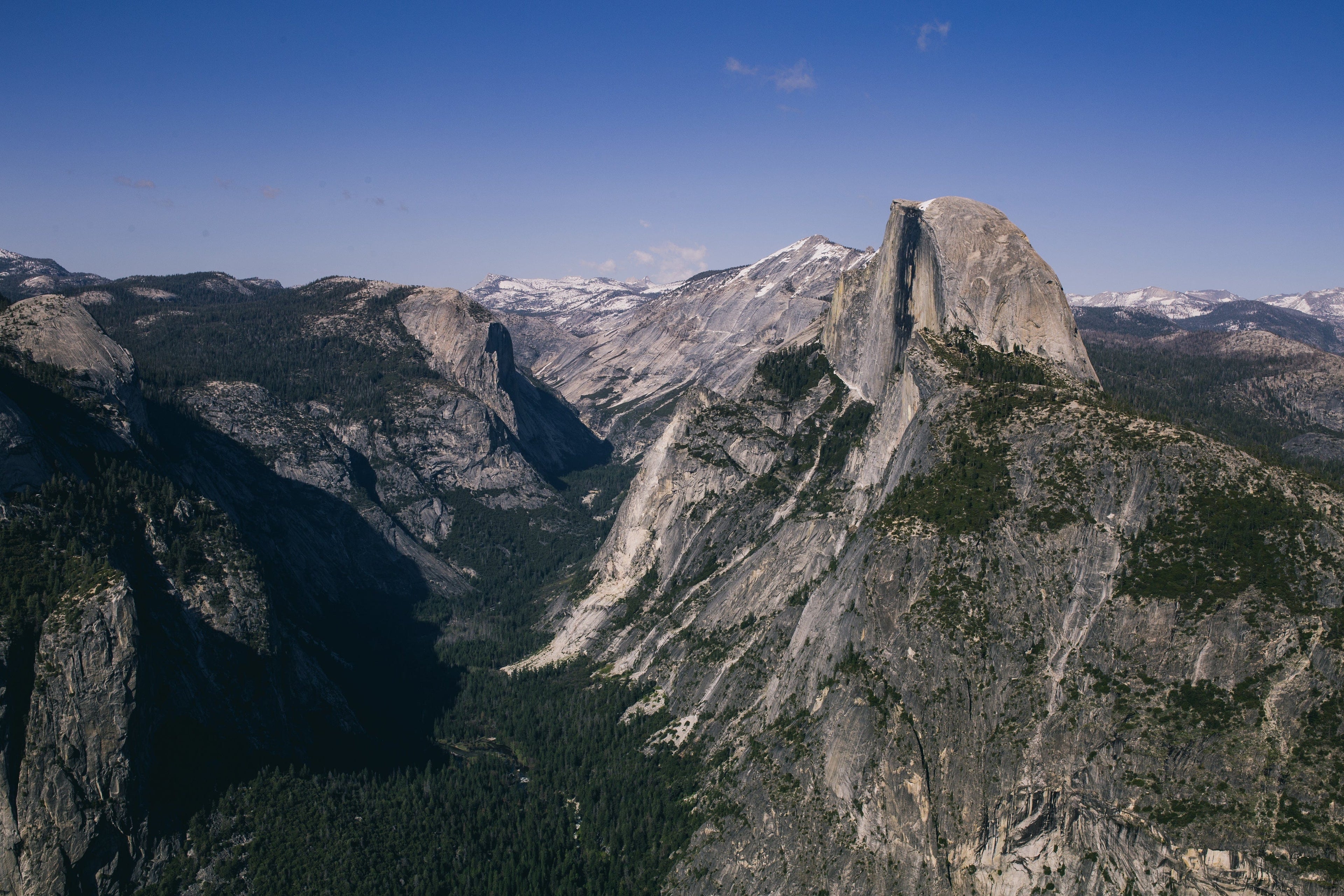 A wide view of Yosemite National Park features Half Dome, a massive granite rock formation, surrounded by forested valleys and distant snow-capped mountains under a clear blue sky.