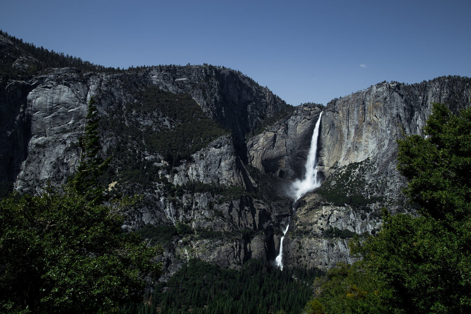 A scenic view of Yosemite Falls cascading down rocky cliffs surrounded by lush green trees and forest, under a clear blue sky.
