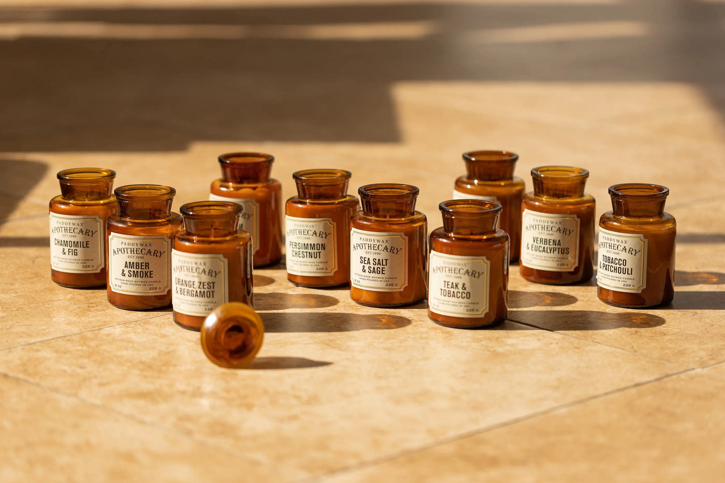 An 8 oz amber glass Verbena + Eucalyptus Apothecary candle is displayed on sunlit tan tiles among other vintage-inspired fragrance bottles. One candle jar rests on its side, highlighting the collection's classic design and soothing scent.