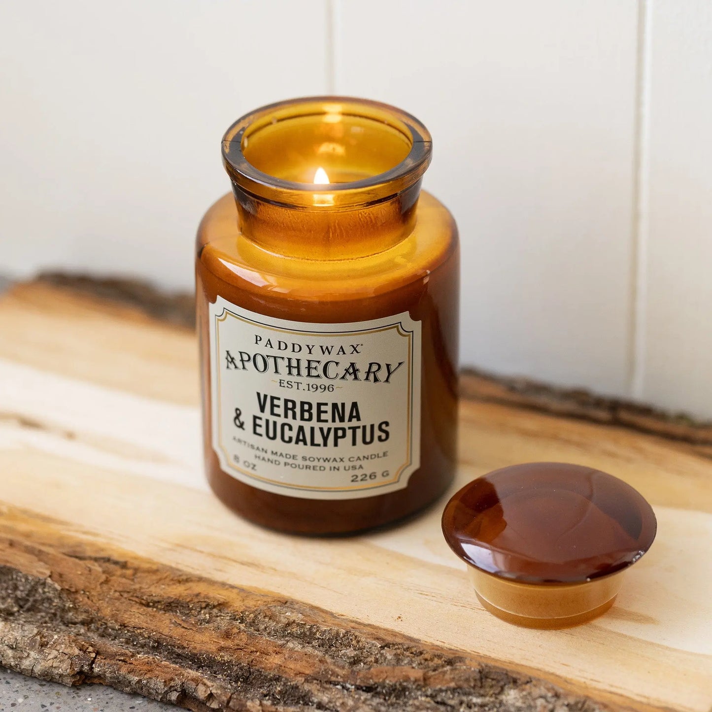 An 8 oz Verbena + Eucalyptus Apothecary Candle in brown amber glass sits on a wooden tray, its vintage-inspired scent evoking nostalgia. The matching lid is beside it, and the jar is partially filled with wax.