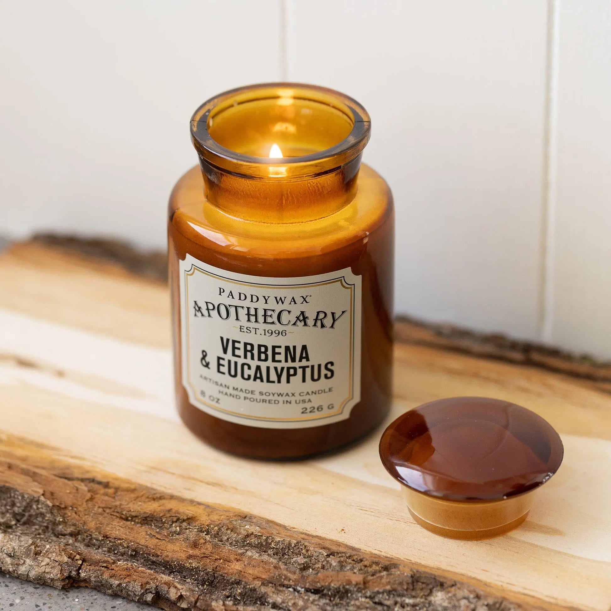 An 8 oz Verbena + Eucalyptus Apothecary Candle in brown amber glass sits on a wooden tray, its vintage-inspired scent evoking nostalgia. The matching lid is beside it, and the jar is partially filled with wax.