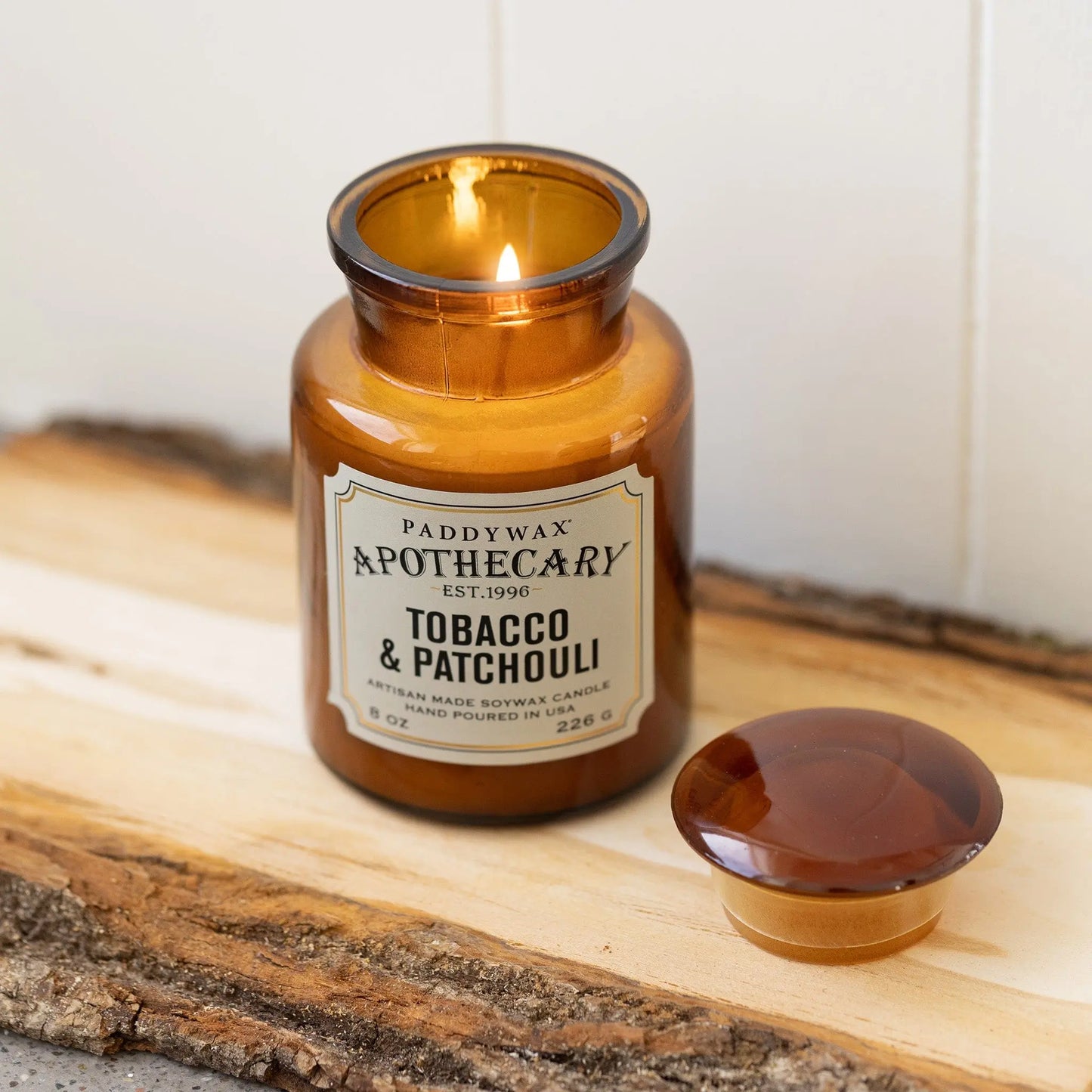 The lit Tobacco + Patchouli - Apothecary 8 oz Candle sits on a wooden surface with its lid nearby, filling the room with a vintage-inspired luxury scent.