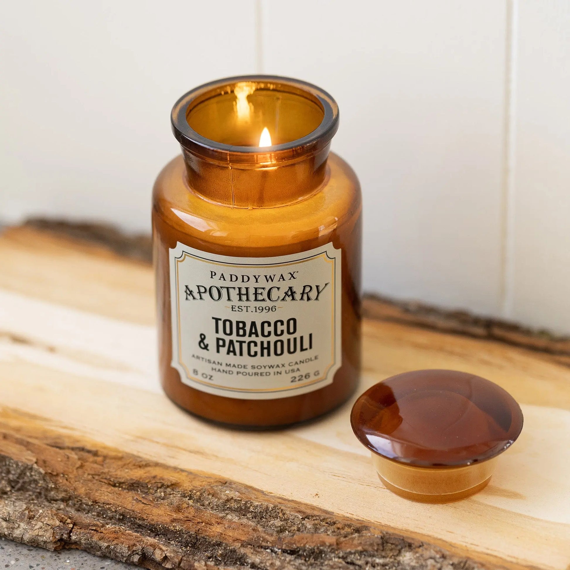 The lit Tobacco + Patchouli - Apothecary 8 oz Candle sits on a wooden surface with its lid nearby, filling the room with a vintage-inspired luxury scent.