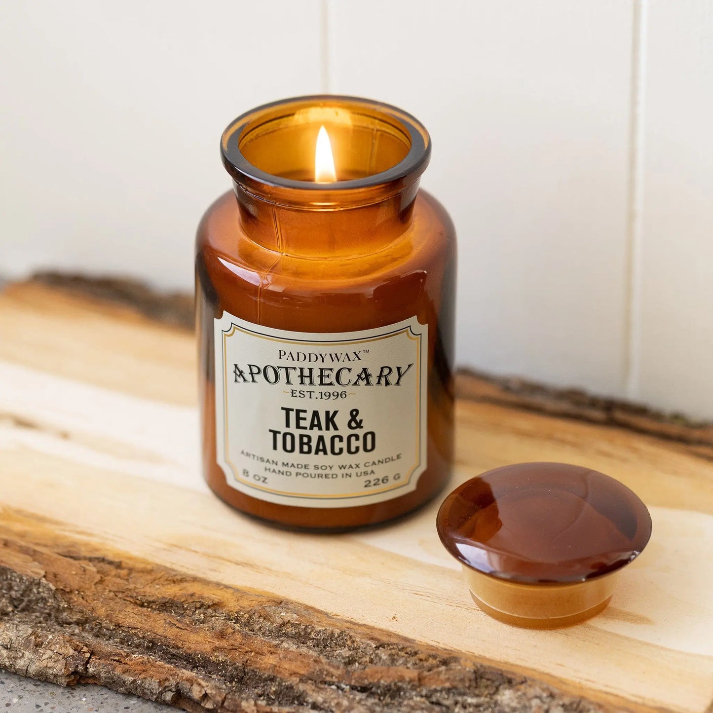8 oz brown apothecary bottle glass vessel with a vintage-style label; white wax; part of the apothecary collection; candle lid off and scented candle burning on wood piece