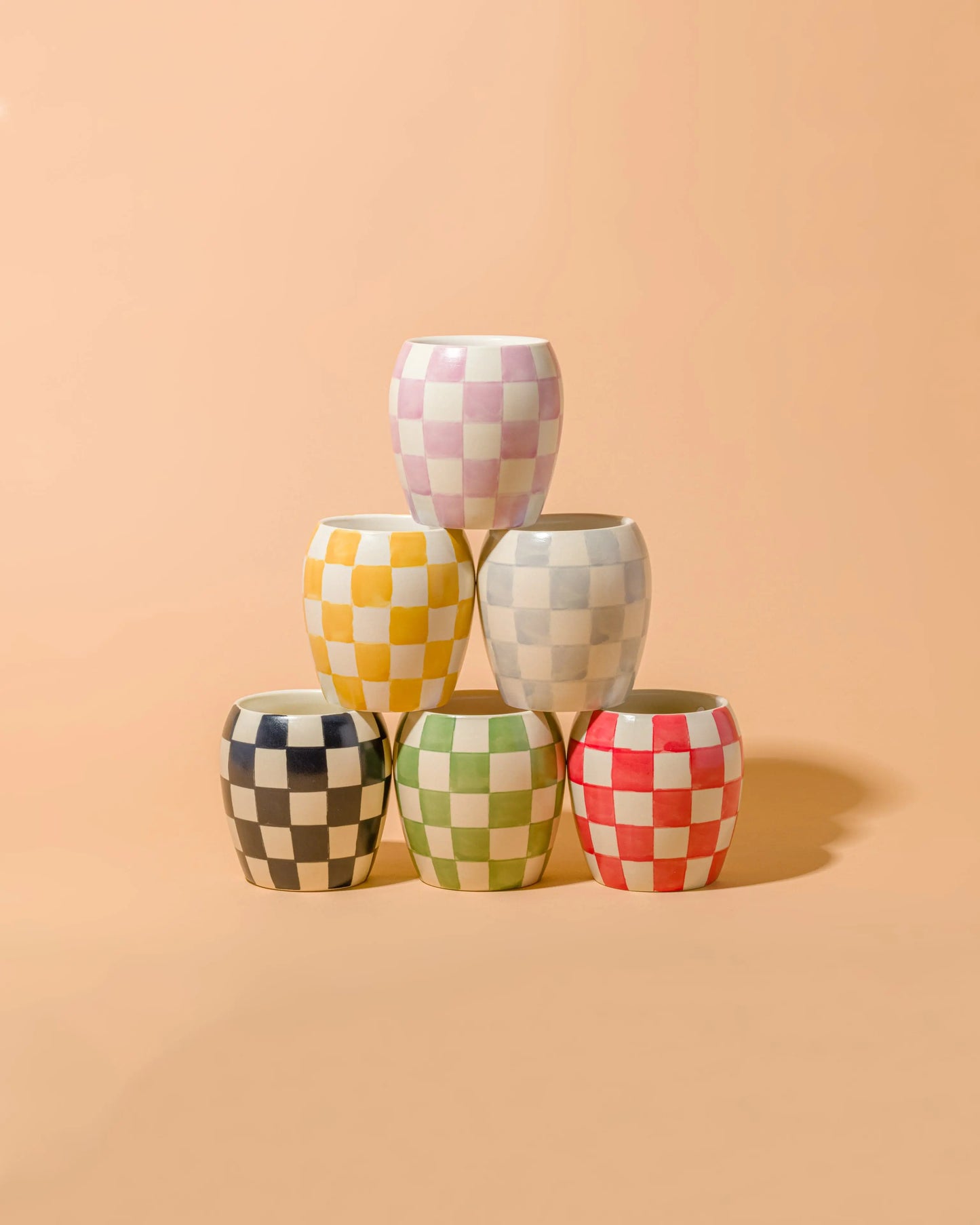 Five Cotton + Teak Checkmate Candles, 11 oz each, with pastel checkerboard ceramic vessels are stacked pyramid-style against a peach background—ideal as reusable decor or unique candle holders.