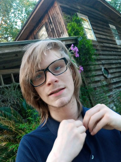 A young person with long blond hair and glasses stands by a rustic wooden house, adjusting their dark shirt. Purple flowers behind their ear and a Black Fig + Olive St. Jude Giveback "Celebrate" 11 oz. Candle help create a warm, inviting scene.