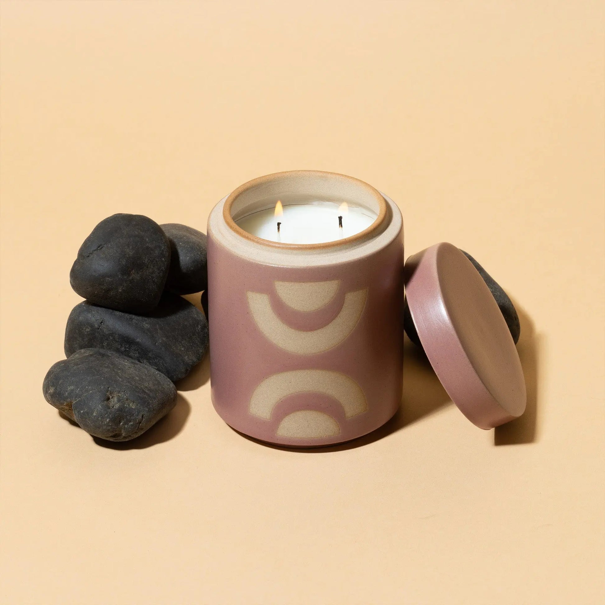 The Mandarin Mango - Form 12 oz Candle, in pink ceramic with geometric designs, sits open beside its lid and smooth black stones on a beige surface, adding a stylish touch to your home decor.