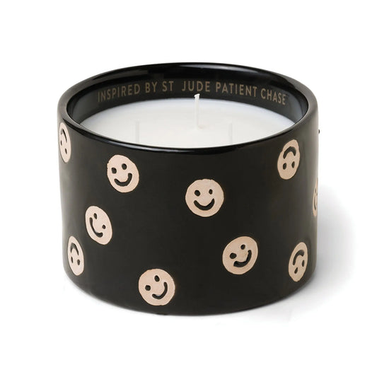 The Black Fig + Olive - St. Jude Giveback "Celebrate" 11 oz. Candle features a black ceramic holder with beige smiley faces, white wax, and INSPIRED BY ST JUDE PATIENT CHASE inscribed inside, making it both meaningful and unique.
