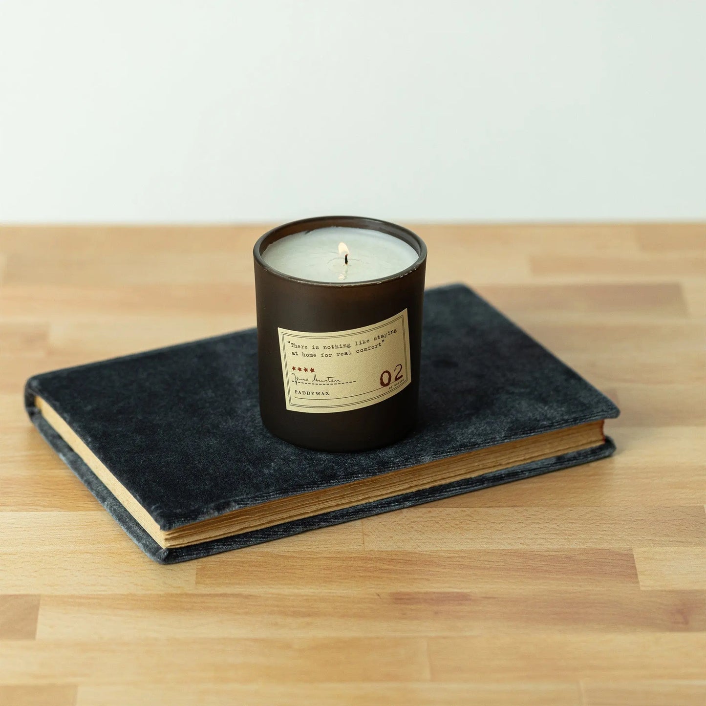 A Jane Austen - Library Candle 6 oz with a label sits on top of a closed dark blue hardcover book on a wooden surface.