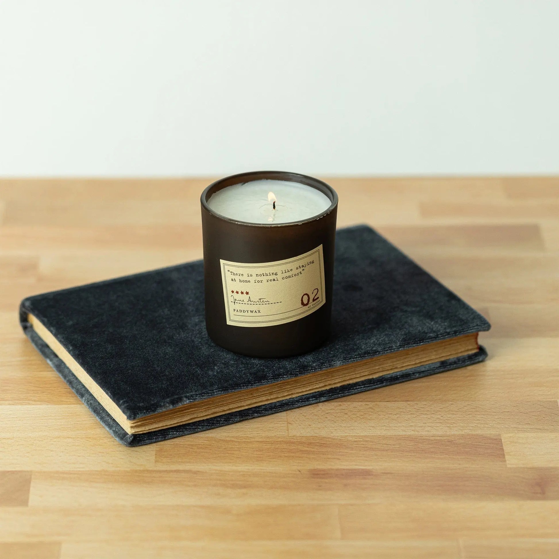 A Jane Austen - Library Candle 6 oz with a label sits on top of a closed dark blue hardcover book on a wooden surface.