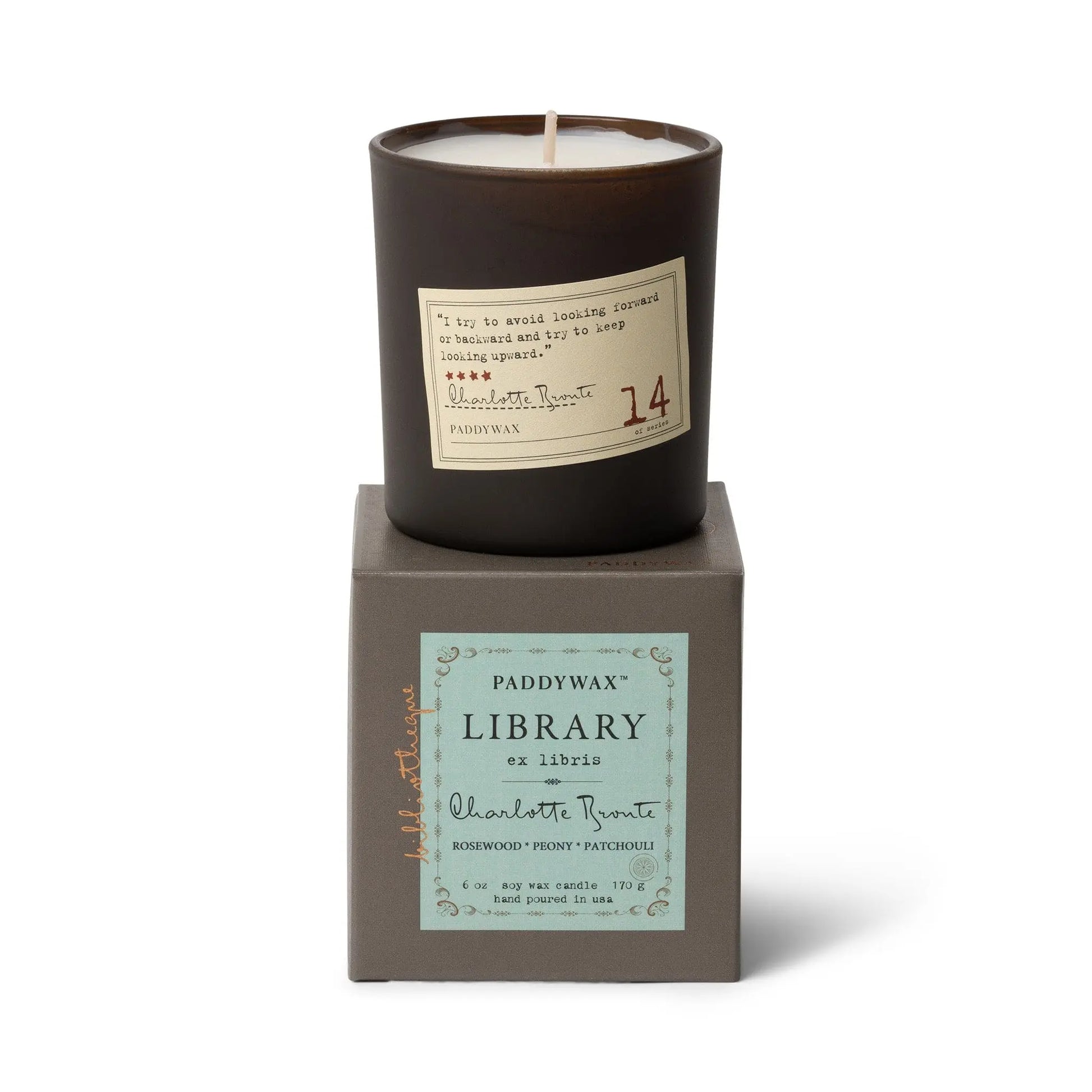 The Charlotte Bronte - Library Candle 6 oz, a dark brown literary candle in a glass jar with rosewood, peony, and patchouli notes, sits atop a gray Paddywax Library Collection box—an ideal fragrance gift for book lovers.