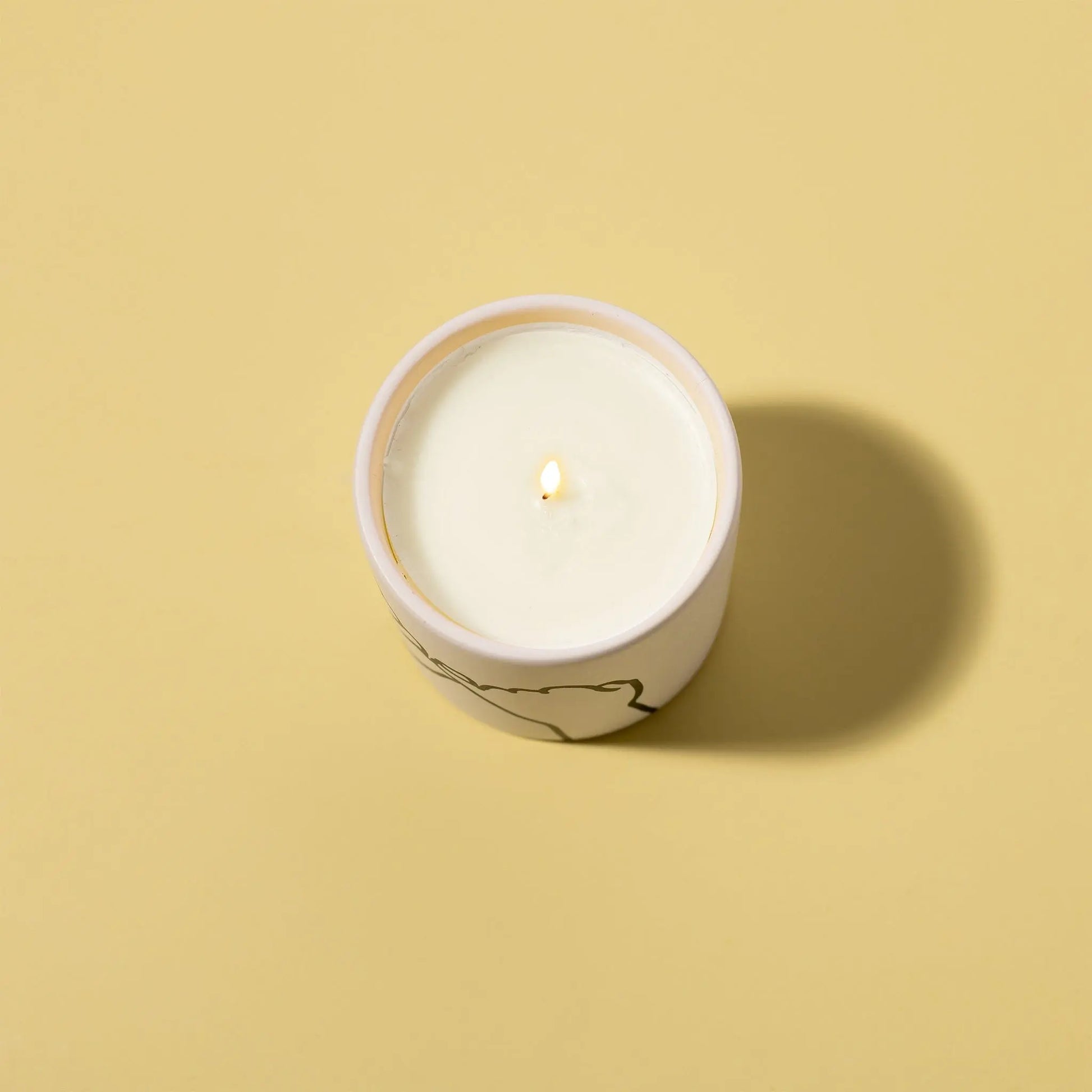 The Wild Fig + Cedar "Pinky Promise" - Impressions 5.75 oz Candle is lit in a round container on a plain beige background, casting a shadow to the right.