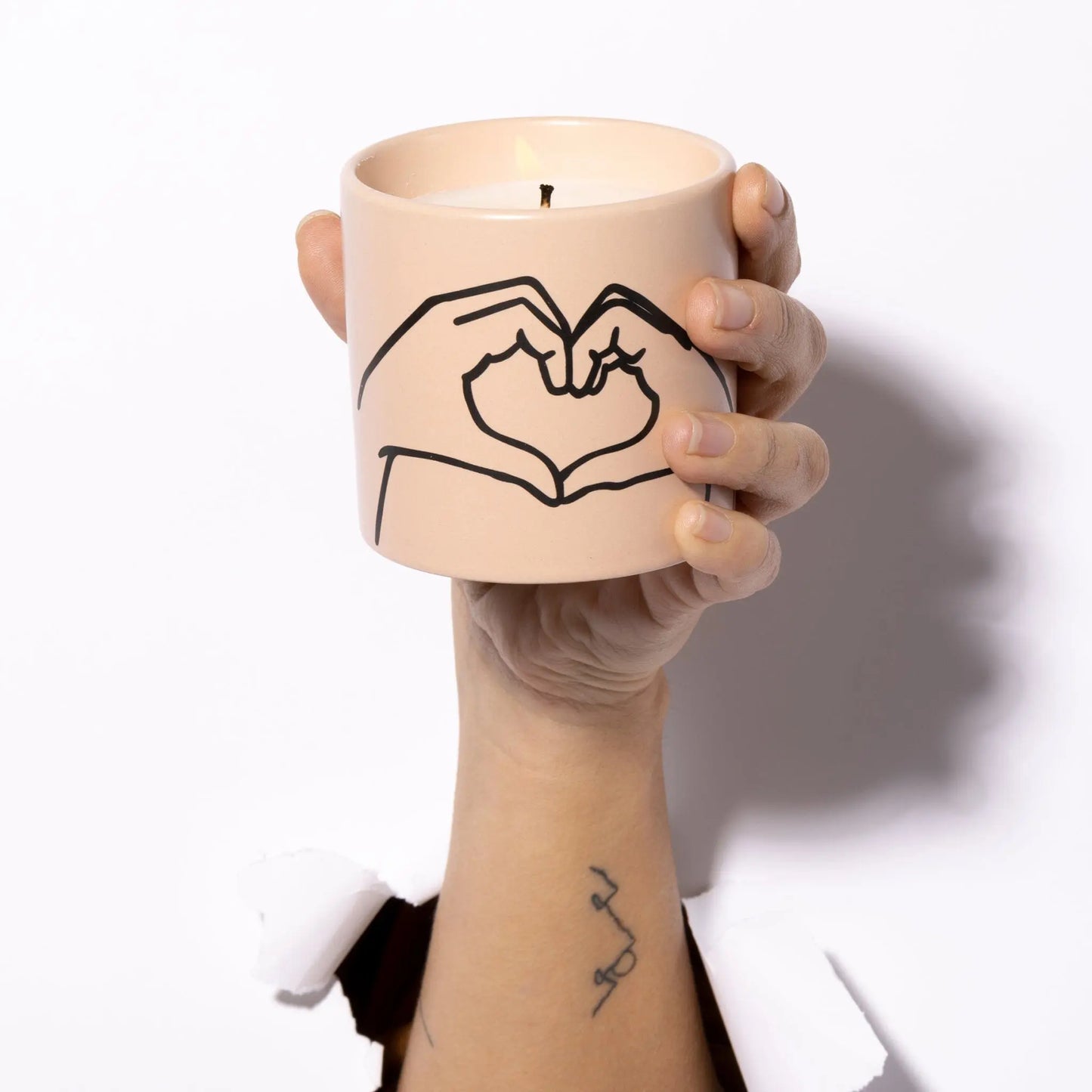 A hand emerges through white paper, holding the Tobacco + Vanilla "Love Ya" Impressions 5.75 oz Candle in a beige ceramic holder adorned with a minimalist line drawing of two hands forming a heart.