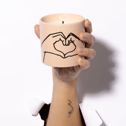A hand emerges through white paper, holding the Tobacco + Vanilla "Love Ya" Impressions 5.75 oz Candle in a beige ceramic holder adorned with a minimalist line drawing of two hands forming a heart.