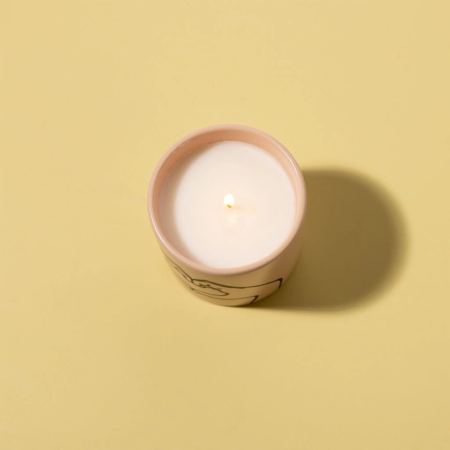 The Tobacco + Vanilla "Love Ya" - Impressions 5.75 oz Candle features a lit white candle in a pink ceramic container on a light yellow surface, casting a soft shadow—ideal for fans of elegant ceramic candles.