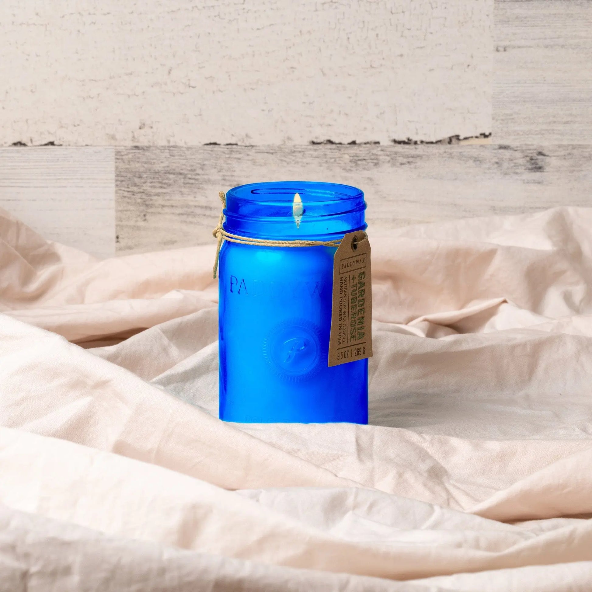 The Gardenia & Tuberose - Relish 9.5 oz Candle, in a lit blue glass jar with a tag, sits on crumpled beige fabric, adding home fragrance to a light wood background.