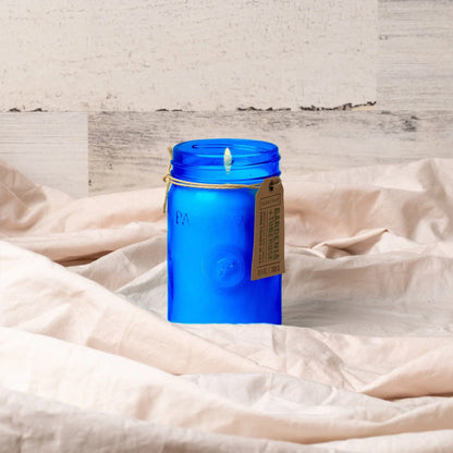 The Gardenia & Tuberose - Relish 9.5 oz Candle, in a lit blue glass jar with a tag, sits on crumpled beige fabric, adding home fragrance to a light wood background.