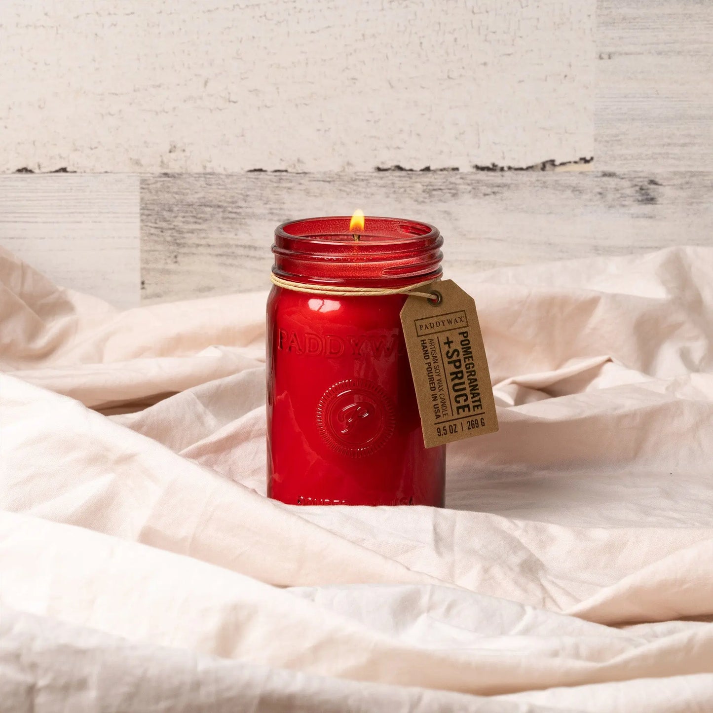 A lit Pomegranate & Spruce - Relish 9.5 oz Candle with a brown tag rests on soft, light-colored fabric against a rustic white wooden background.
