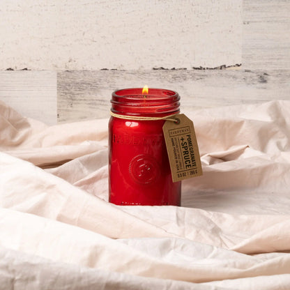 A lit Pomegranate & Spruce - Relish 9.5 oz Candle with a brown tag rests on soft, light-colored fabric against a rustic white wooden background.