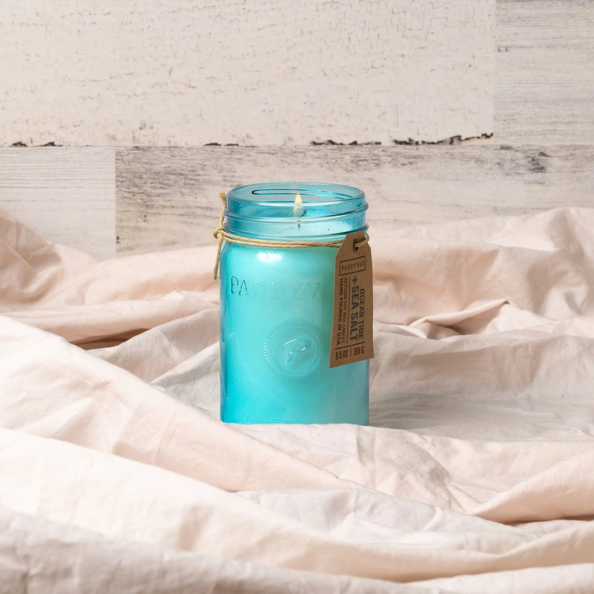 The Ocean Tide & Sea Salt - Relish 9.5 oz Candle, featuring a blue hue in an upcycled glass jar with a tan paper tag, is displayed on beige fabric with a light wood backdrop.