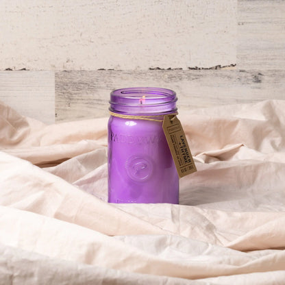 The Lavender + Thyme - Relish 9.5 oz Candle is a purple scented candle in a mason jar with a label, displayed on beige fabric against a light wood background.