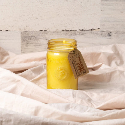 The Fresh Meyer Lemon - Relish 9.5 oz Candle, in a glass jar with a brown label, rests on crumpled beige fabric against a light wooden background, spreading its delightful lemon fragrance.