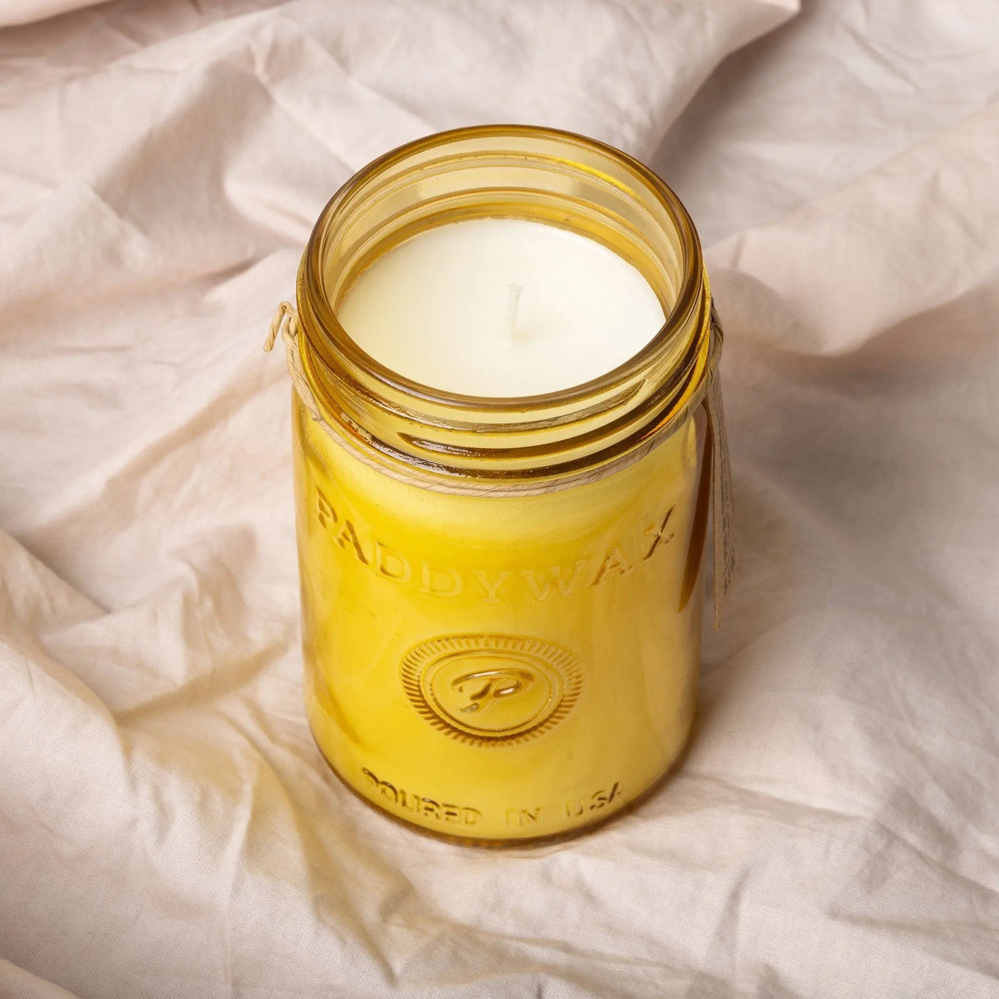 A Fresh Meyer Lemon - Relish 9.5 oz Candle in a yellow Paddywax glass jar with a white wick rests on light, crumpled fabric, ready to brighten your space with its citrus fragrance.