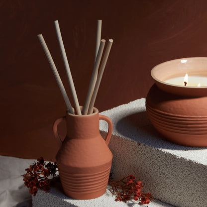 A Santorini 4 fl oz. Diffuser in Raw Clay & Pear, featuring a terracotta vase with reed sticks and red flowers, sits beside a lit candle on a textured white surface.
