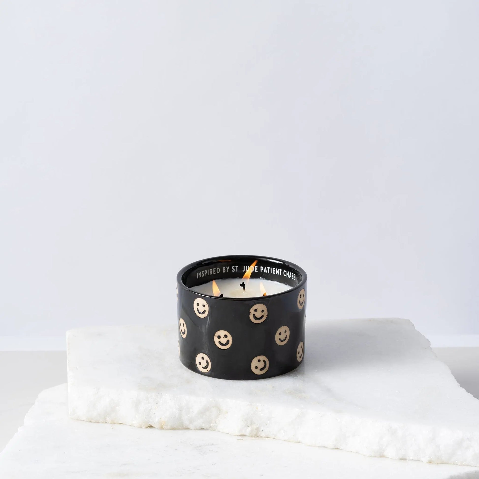 The Black Fig + Olive - St. Jude Giveback "Celebrate" 11 oz. Candle is lit, featuring black ceramic with beige smiley faces, and sits on white marble slabs against a plain light background.