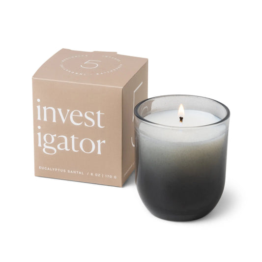 A gray ombre glass Enneagram #5 Investigator 5 oz Candle in eucalyptus santal scent burns beside its beige box, featuring modern minimalist typography—the ideal gift for those who value unique, thoughtful design.